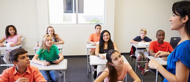 Photo of a middle school classroom with the teacher talking to students at the front of the class.