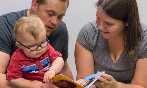 parents reading a book to a baby who is wearing glasses