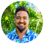 Photo of smiling Native Hawaiian man with dark hair and beard, in front of green hedges.