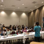 People sitting on long tables in a room facing a man in the front of the room