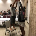 Two people stretching their arms up to hold the top of their tall straw tower. One person is standing on a chair.