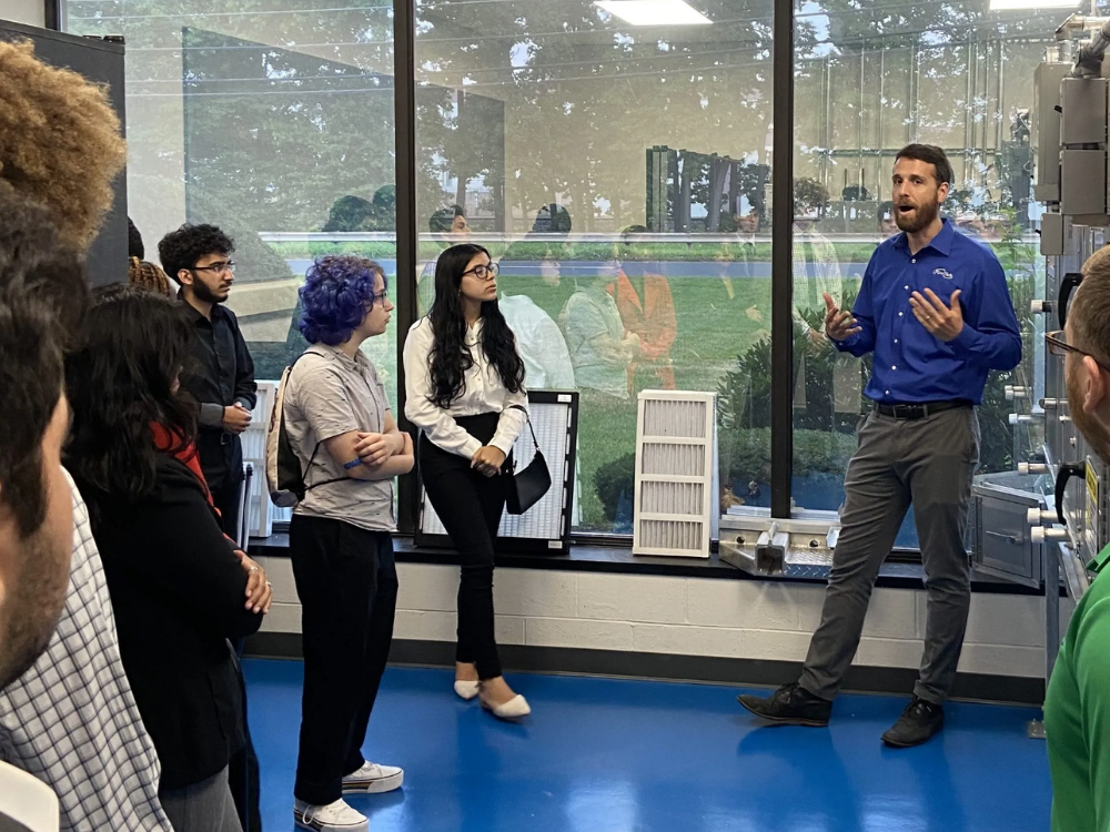 A lecturer is standing and giving a speech to a group of surrounding students.